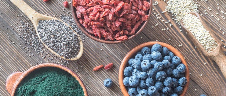 A flat lay of different superfoods in bowls against a wooden table, including goji berries, blueberries and chia seeds.