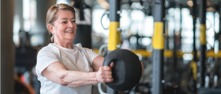 A woman over 50 strength training with a weighted medicine ball in a gym.