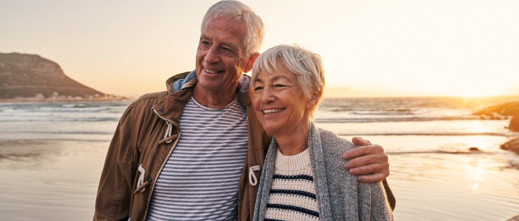 An elderly couple smiling and walking together on a beach at sunset because they passed their general medical check up.