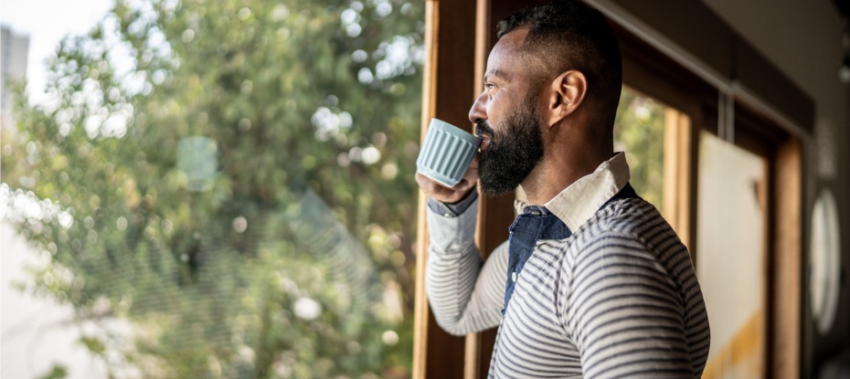 A man looking out of his window, sipping on a drink from a cup