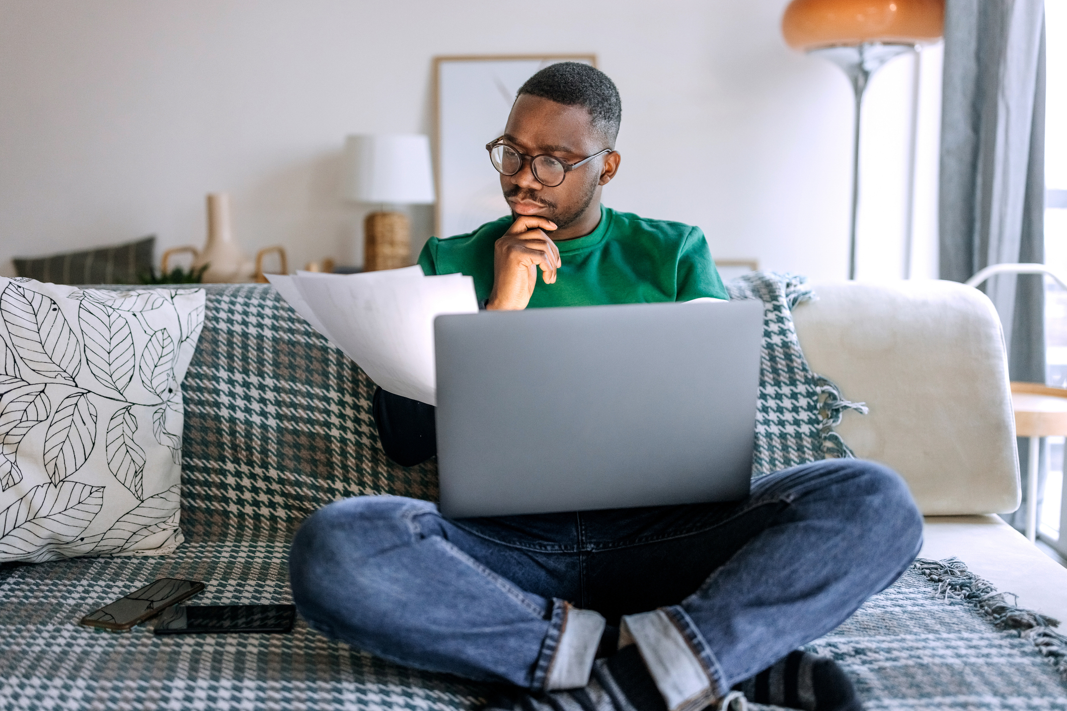 A man in a green jumper and blue denim jeans is sitting on a couch pondering Health Insurance for Visitor Visa 600.