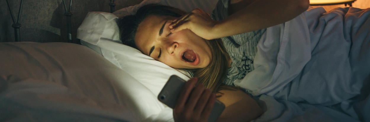 woman laying in bed yawning while on her phone 