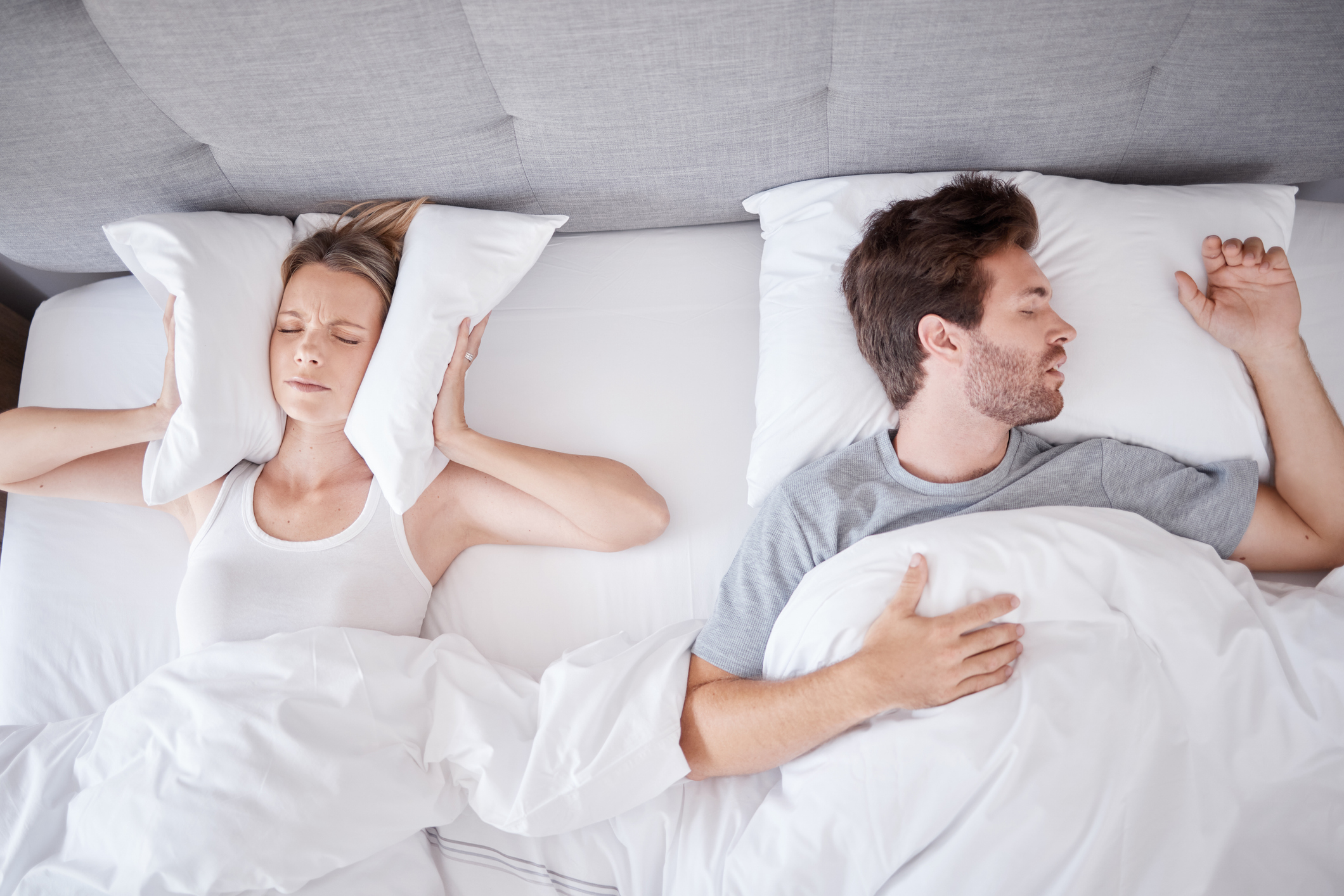 A couple lying in bed, with the woman covering her ears with a pillow, possibly due to her partner's loud snoring caused by sleep apnea