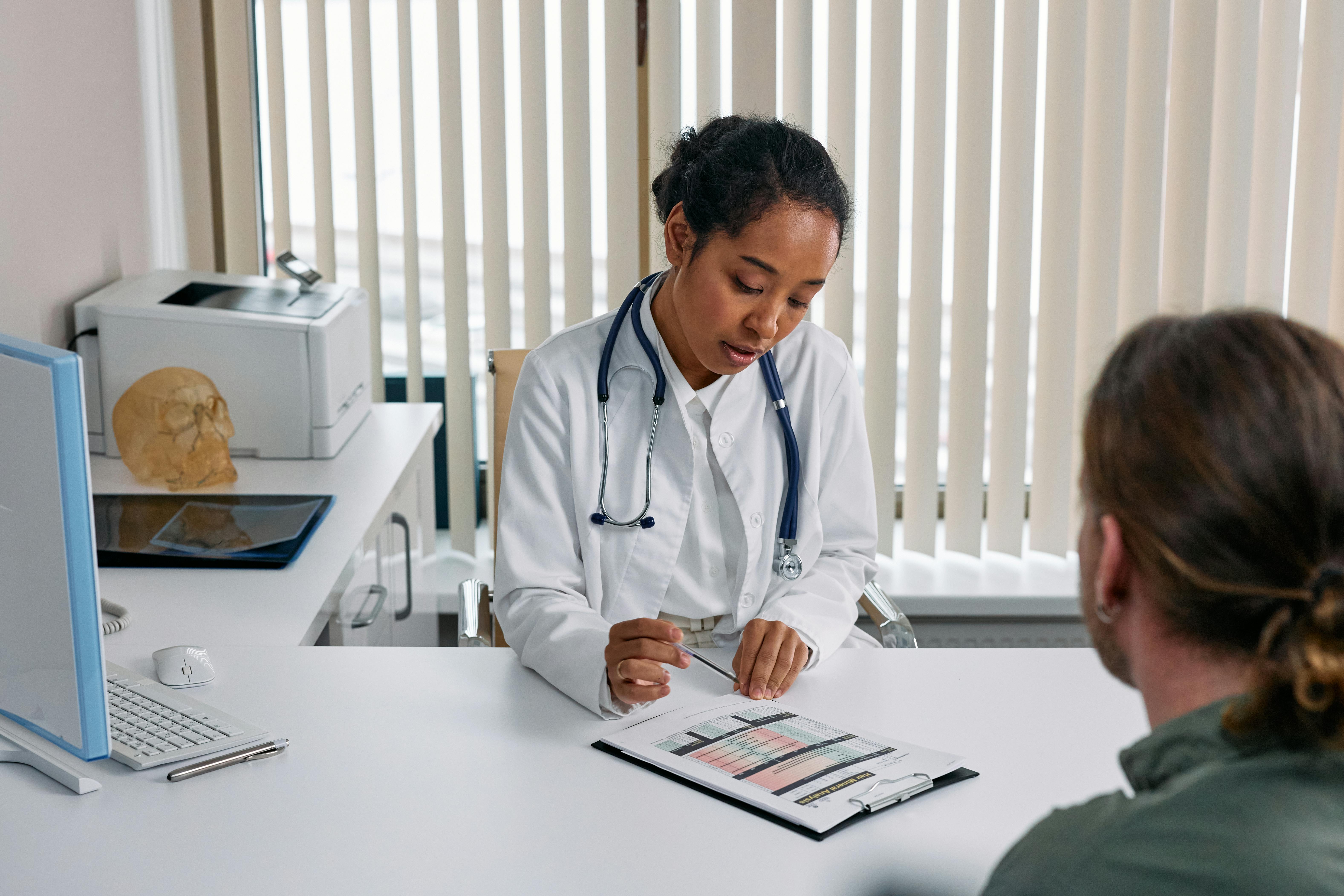 A doctor in a white coat sits with a patient in a medical office, discussing pancreatic cancer. The doctor holds a clipboard with medical records, and the patient listens attentively in a calm, professional setting.